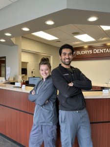 Two smiling dental professionals standing back-to-back with arms crossed in front of a reception desk labeled Budrys Dental