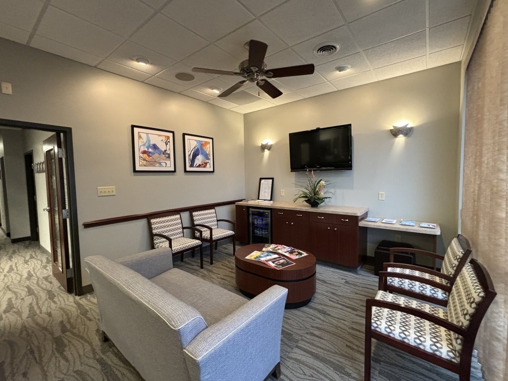 Waiting room lounge with light grey sofa, patterned armchairs, coffee table with magazines, and wall-mounted television
