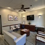 Waiting room lounge with light grey sofa, patterned armchairs, coffee table with magazines, and wall-mounted television