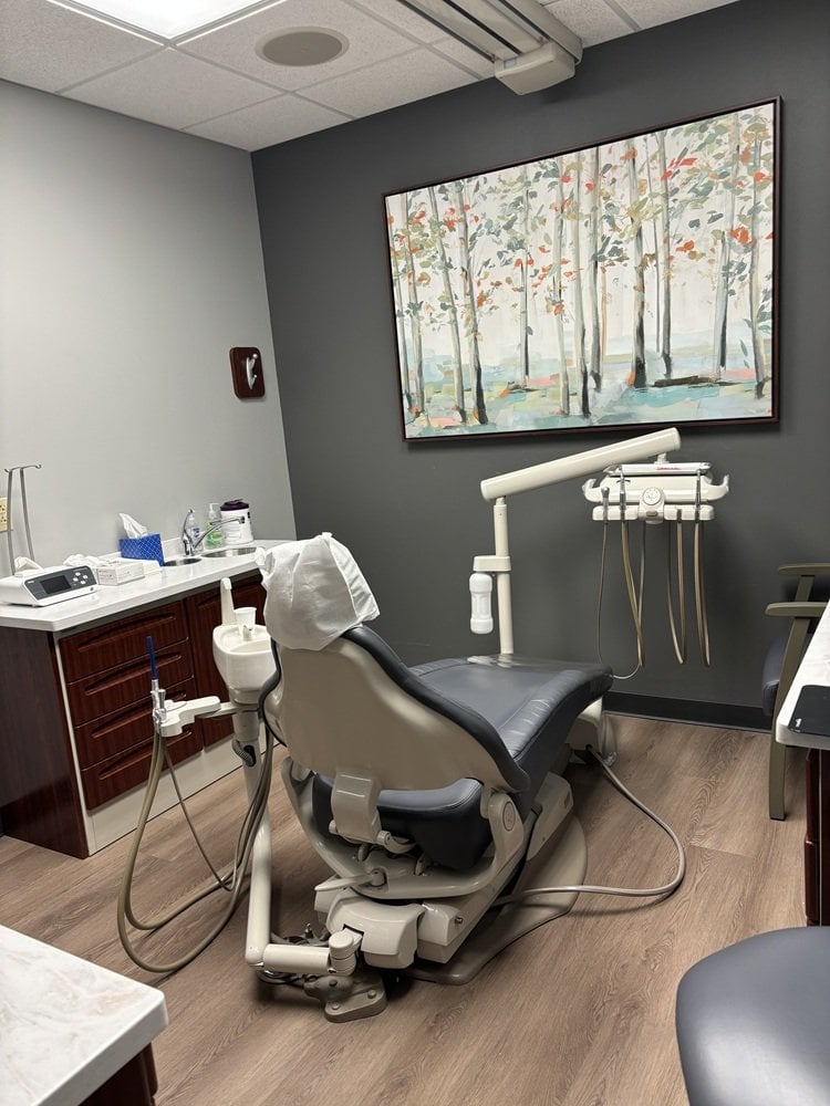 Dental treatment room with a central patient chair, overhead lamp, instrument tray, side cabinets, and framed wall art depicting a forest scene