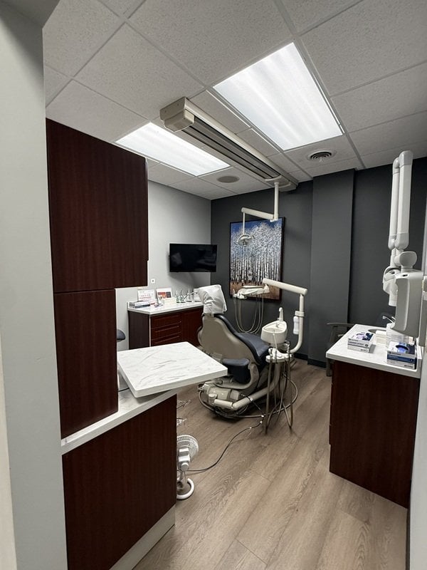 Modern dental operatory featuring a gray patient chair, dark wood cabinetry, overhead light fixture, and framed forest artwork on a gray wal