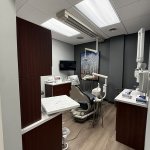 Modern dental operatory featuring a gray patient chair, dark wood cabinetry, overhead light fixture, and framed forest artwork on a gray wal