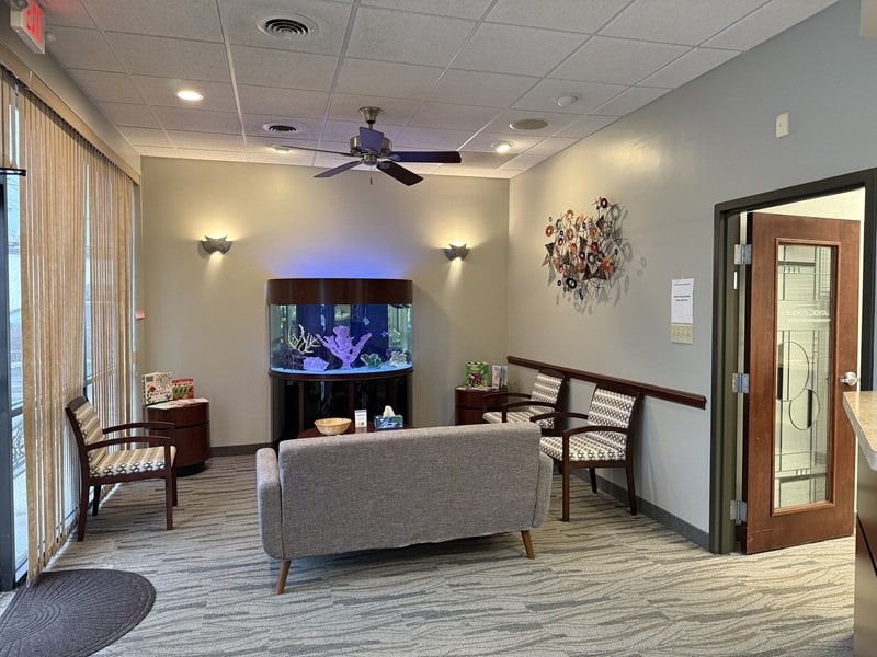 Dental office waiting area featuring a large cylindrical aquarium, grey sofa, accent chairs, and a ceiling fan
