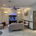 Dental office waiting area featuring a large cylindrical aquarium, grey sofa, accent chairs, and a ceiling fan