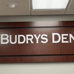 Wall sign displaying BUDRYS DENTAL with a toothbrush logo on a dark wood background